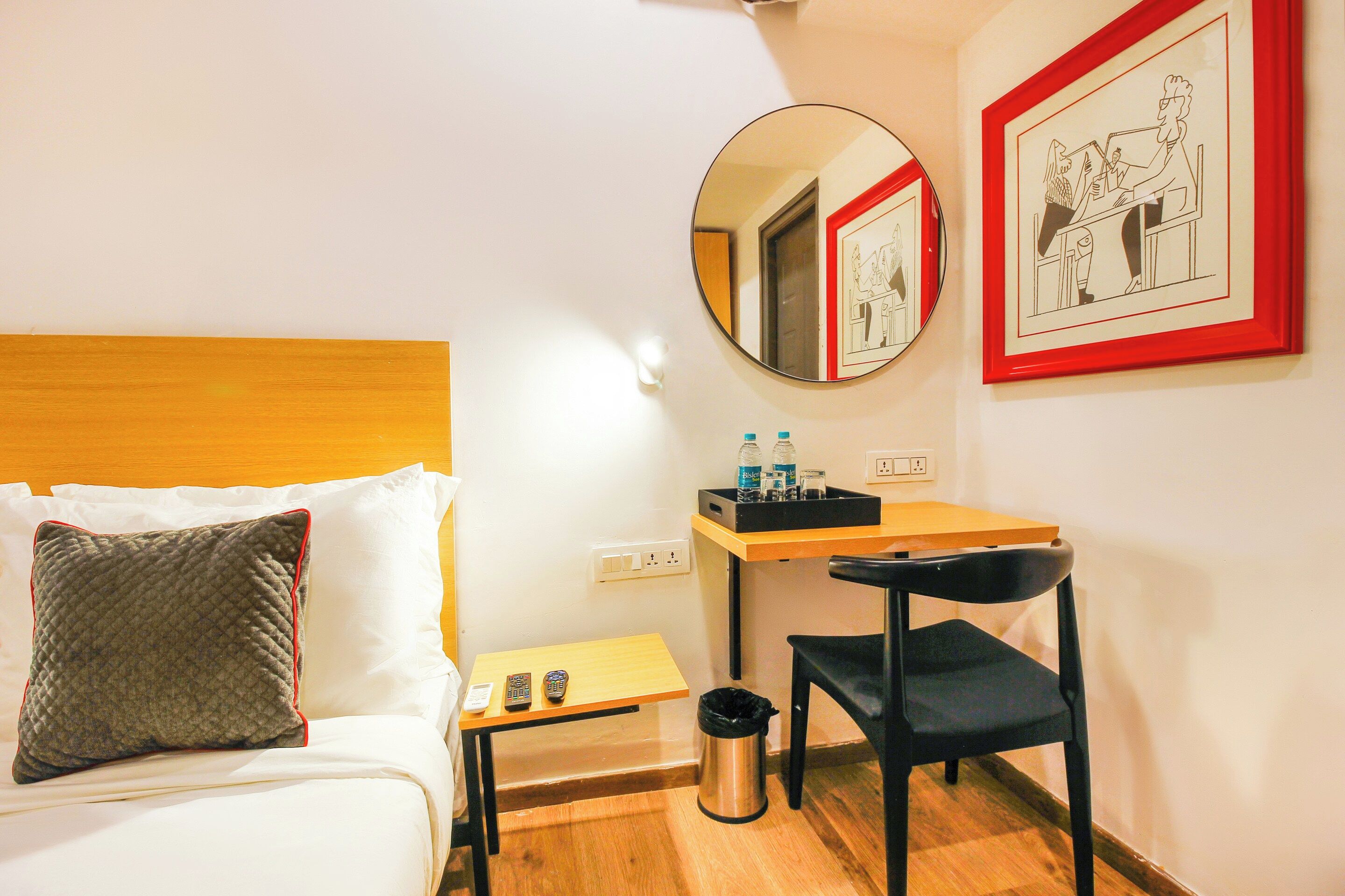 Ein Hotelzimmer mit einem Bett, einem Schreibtisch, einem Stuhl, einem Mülleimer, einem Spiegel und zwei gerahmten Bildern an der Wand.