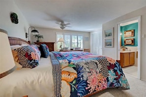 Ein Schlafzimmer mit einem großen Bett, einem Deckenventilator, einem Schreibtisch mit Lampe und Blick ins Freie durch ein Fenster.