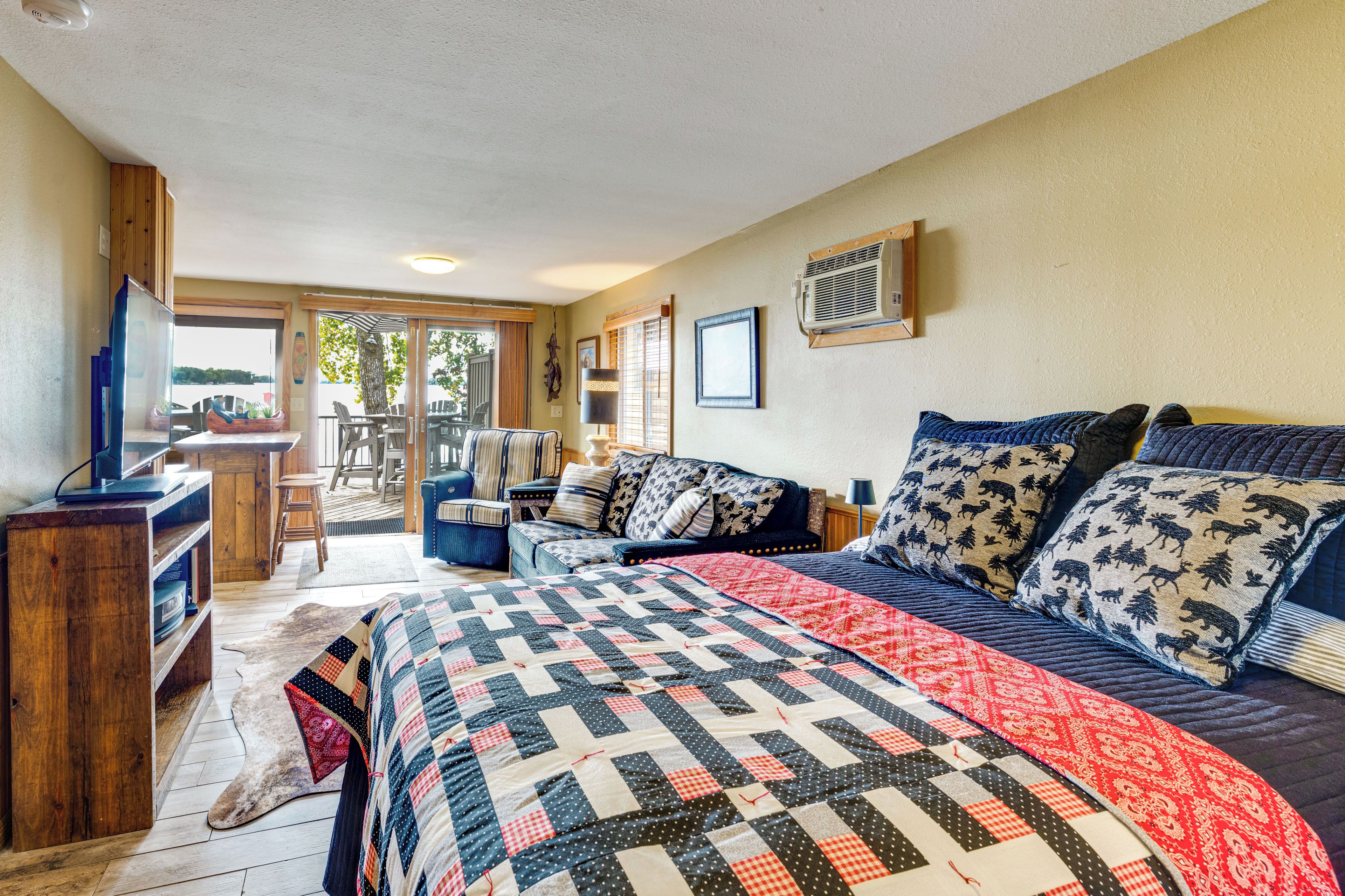 A bedroom with a large bed, a desk, and a chair. There is a window with curtains, a ceiling fan, and a wall-mounted air conditioner.