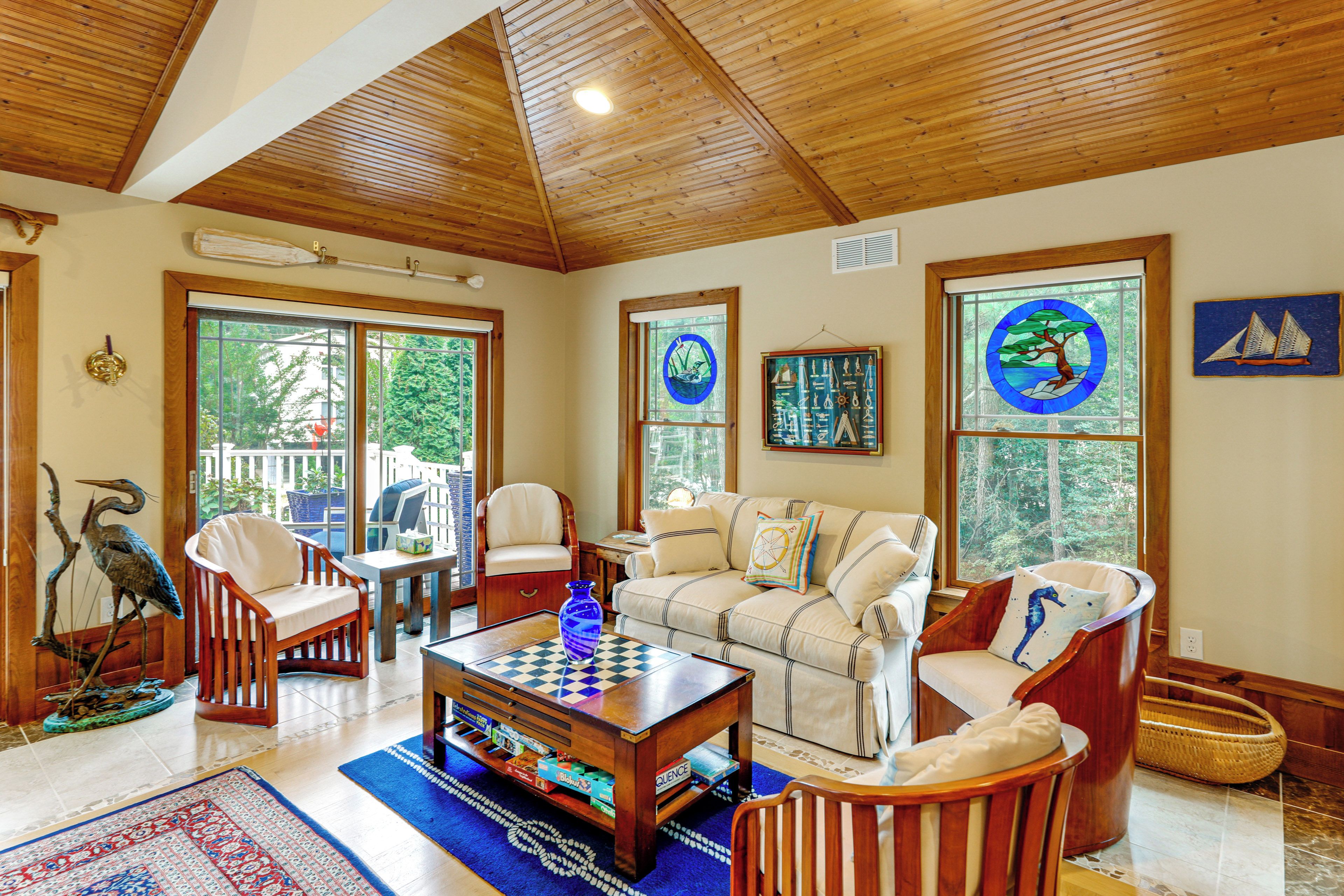 A spacious living room with a high ceiling, wooden beams, a fireplace, a large window with a view of trees, and a cozy seating area with a coffee table and chairs.