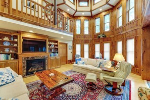 A spacious living room with a fireplace, wooden furniture, and a large window overlooking a scenic view. - Walk to Beach! 7BR Group Retreat in Bethany Beach (Bethany Beach)