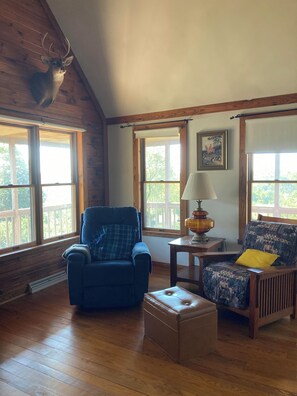 A room with wooden floors, a blue armchair, a patterned armchair, a small table with a lamp, and a deer head mounted on the wall. - 2BR + spacious loft. Rich Hill Retreat offers spectacular mountain view. (Connellsville)