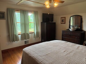 Un dormitorio con cama, cómoda, televisor sobre un mueble, ventana con cortinas y ventilador de techo.