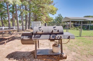 A portable barbecue grill with multiple burners, a gas tank, and a metal table in a grassy outdoor area with trees and a building in the background.