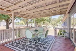 A covered outdoor patio with a table and chairs, a patterned rug, and a view of trees and a fence in the background.