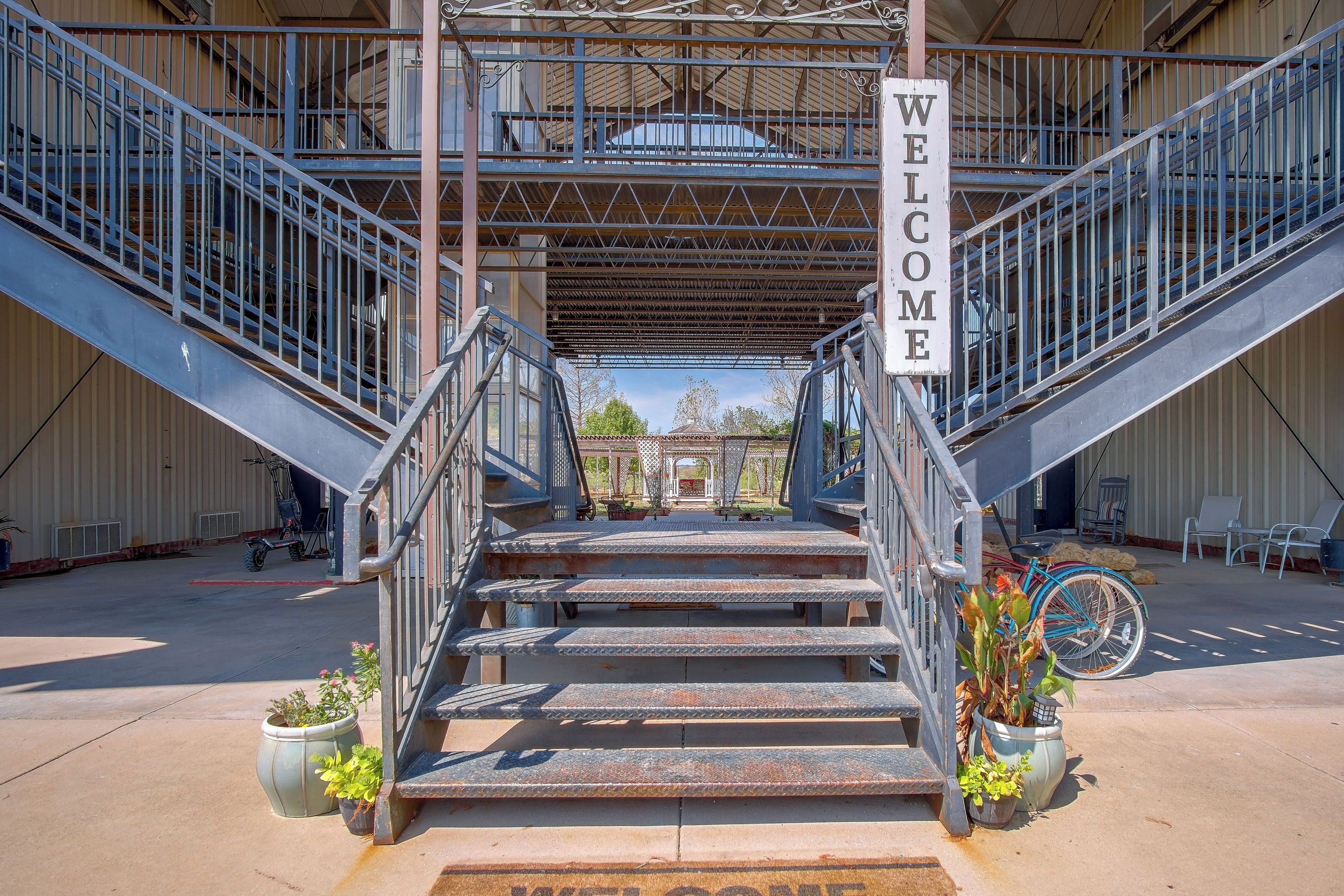 Una escalera metálica conduce a una zona cubierta con un letrero que dice 'WELCOME', rodeada de macetas con plantas y asientos exteriores.