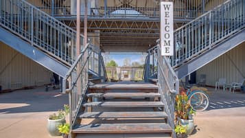 Una escalera metálica conduce a una zona cubierta con un letrero que dice 'WELCOME', rodeada de macetas con plantas y asientos exteriores.