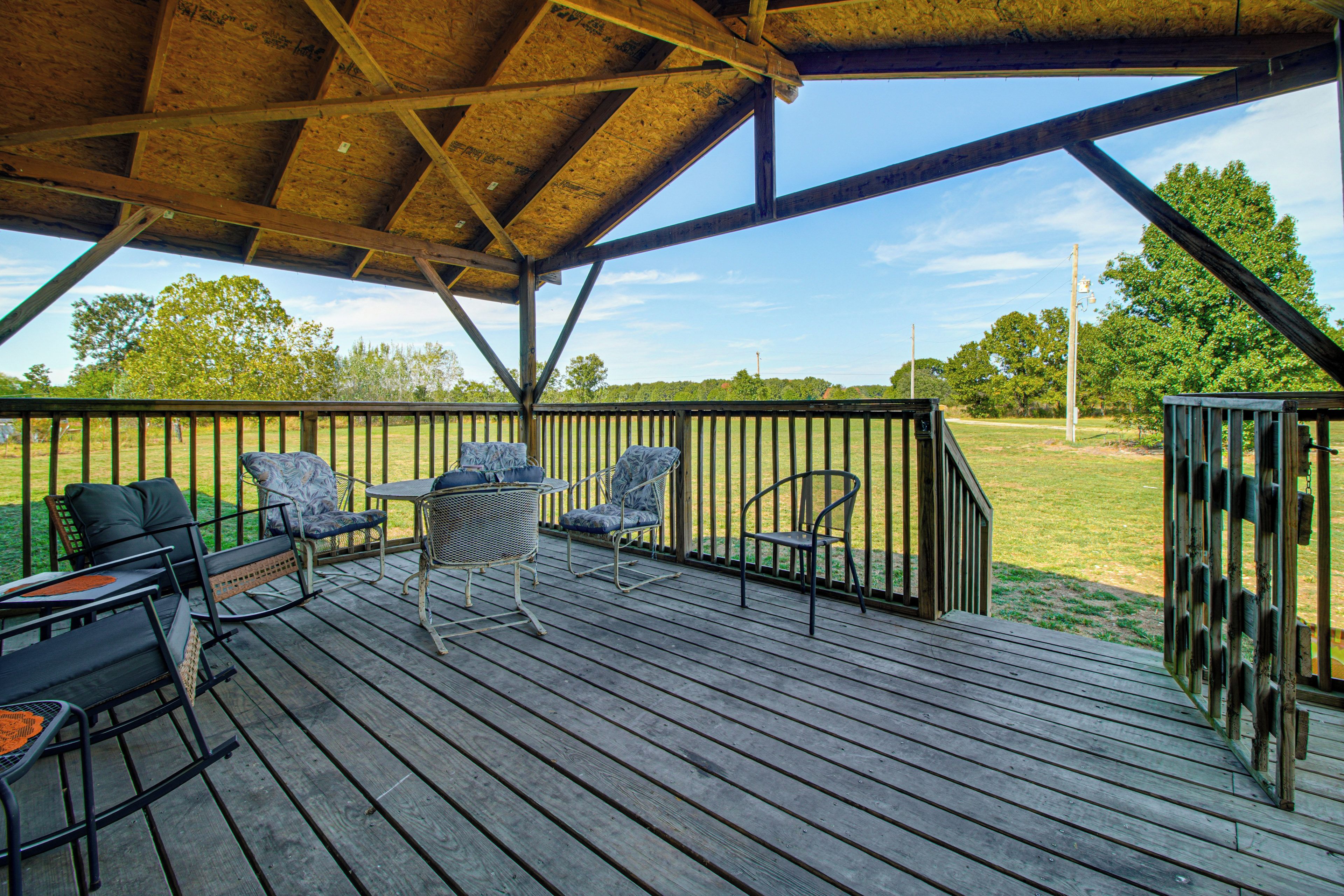 A covered outdoor deck with wooden flooring and metal railings, featuring outdoor furniture and a view of a grassy field.