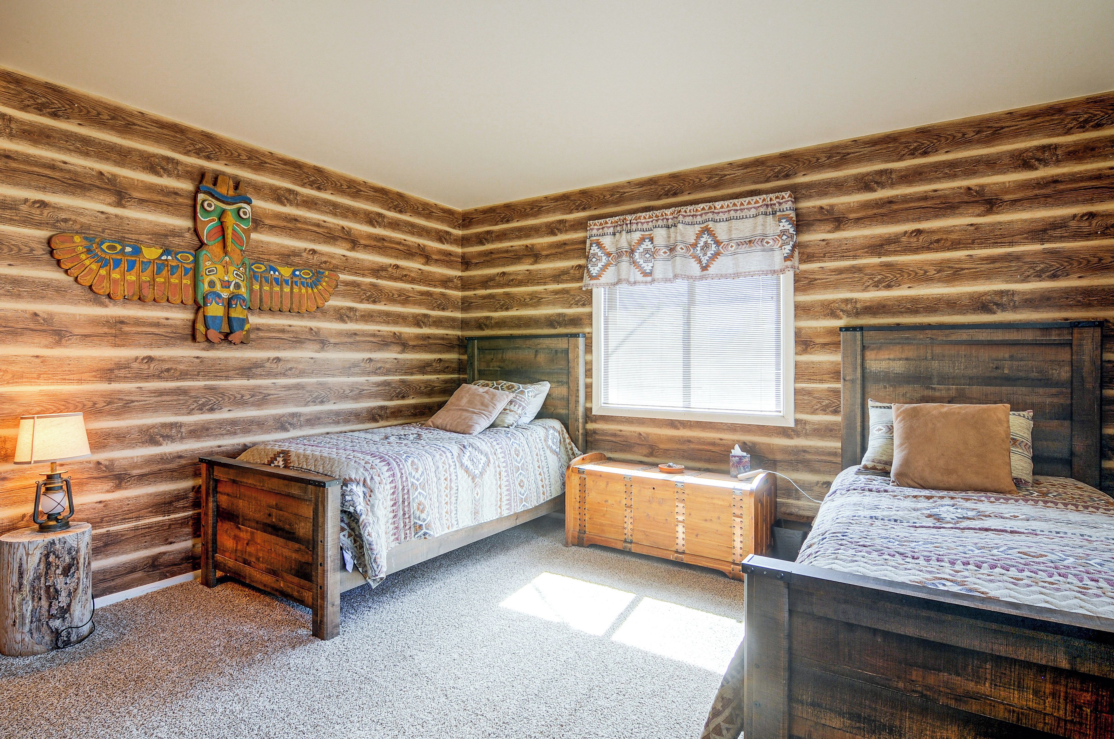 A log cabin-style bedroom with two beds, a wooden headboard, a window with blinds, and a decorative wall piece.