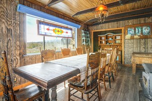 A rustic dining room with a large wooden table and chairs, a stained glass window, and wooden shelves with decorative items.