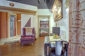 Quarto com piso de madeira, uma poltrona roxa, uma mesa de jantar com cadeiras, uma TV na parede e uma estante com utensílios de cozinha.