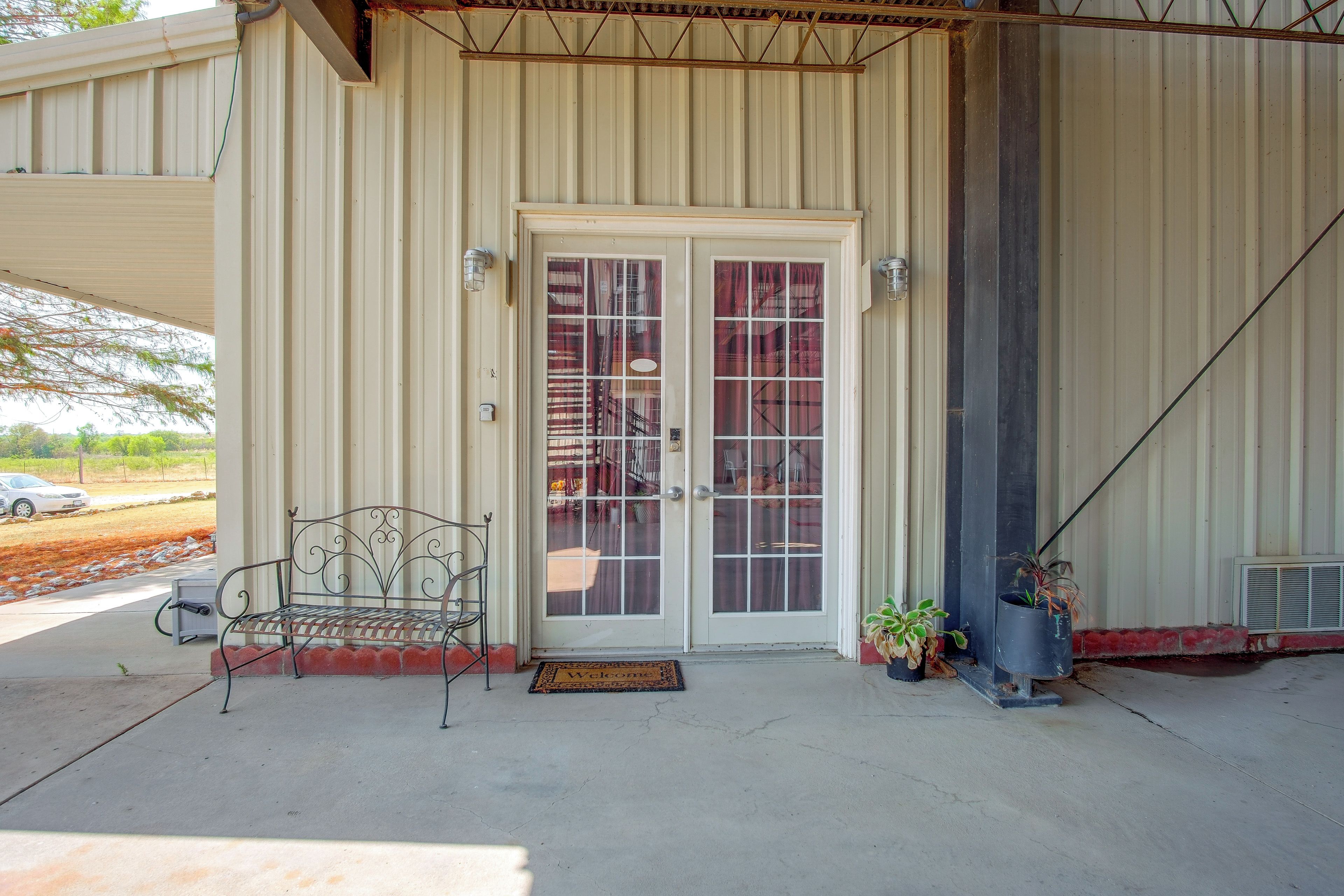A building entrance with a metal bench, a welcome mat, and potted plants.