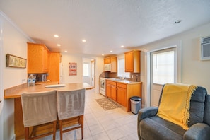 A compact kitchen with wooden cabinets, a dining area with two chairs, a sofa with a yellow throw, and a window with blinds. - Naples Duplex Bundle By Shopping & Beaches (Naples)