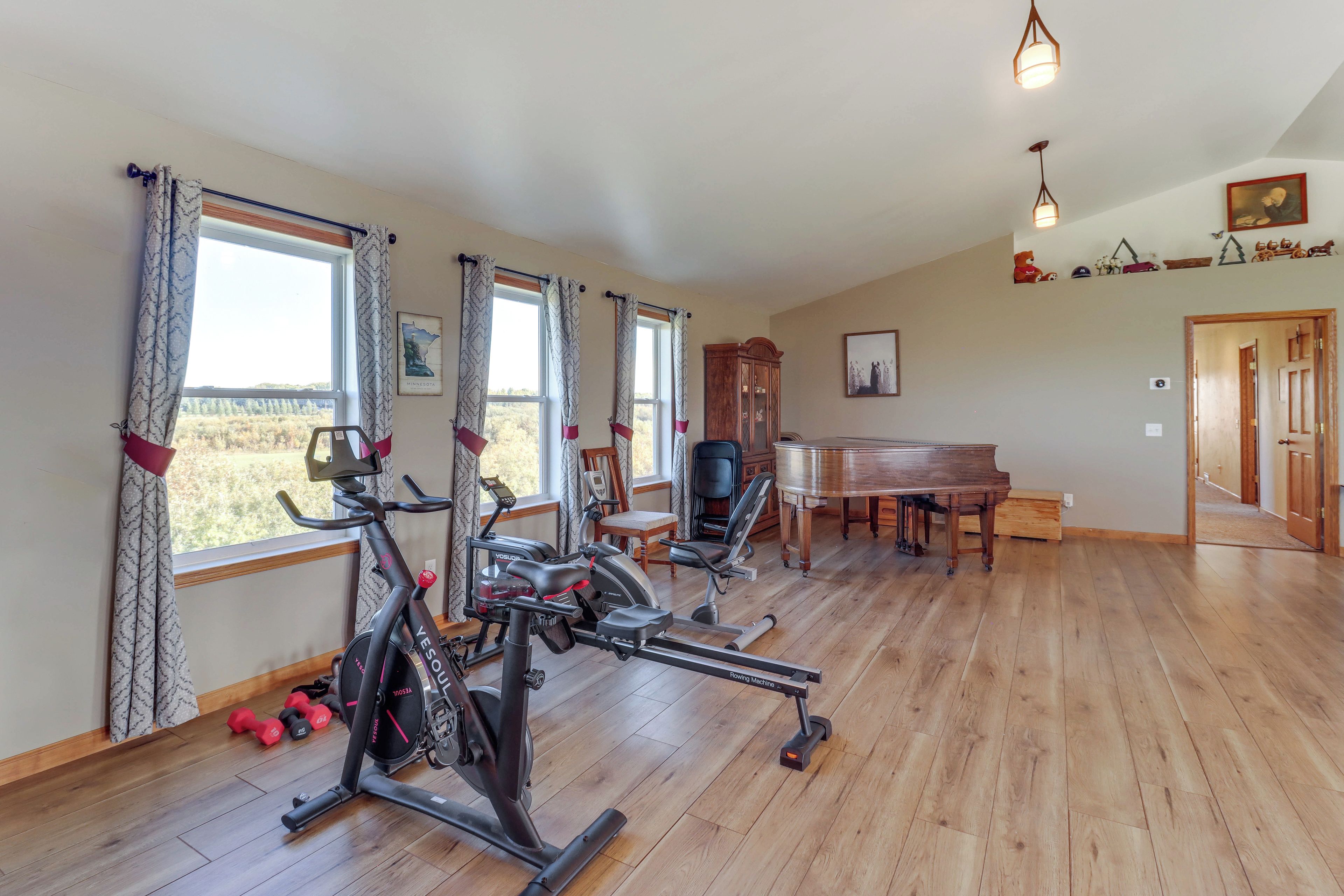 A spacious room with a wooden floor, a piano, exercise equipment, and large windows with curtains.