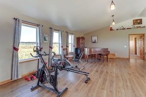 Una habitación amplia con suelo de madera, un piano, equipo de ejercicio y ventanas grandes con cortinas.