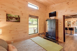 A wooden cabin interior with a bed, a cabinet, a television, and a kitchen area.
