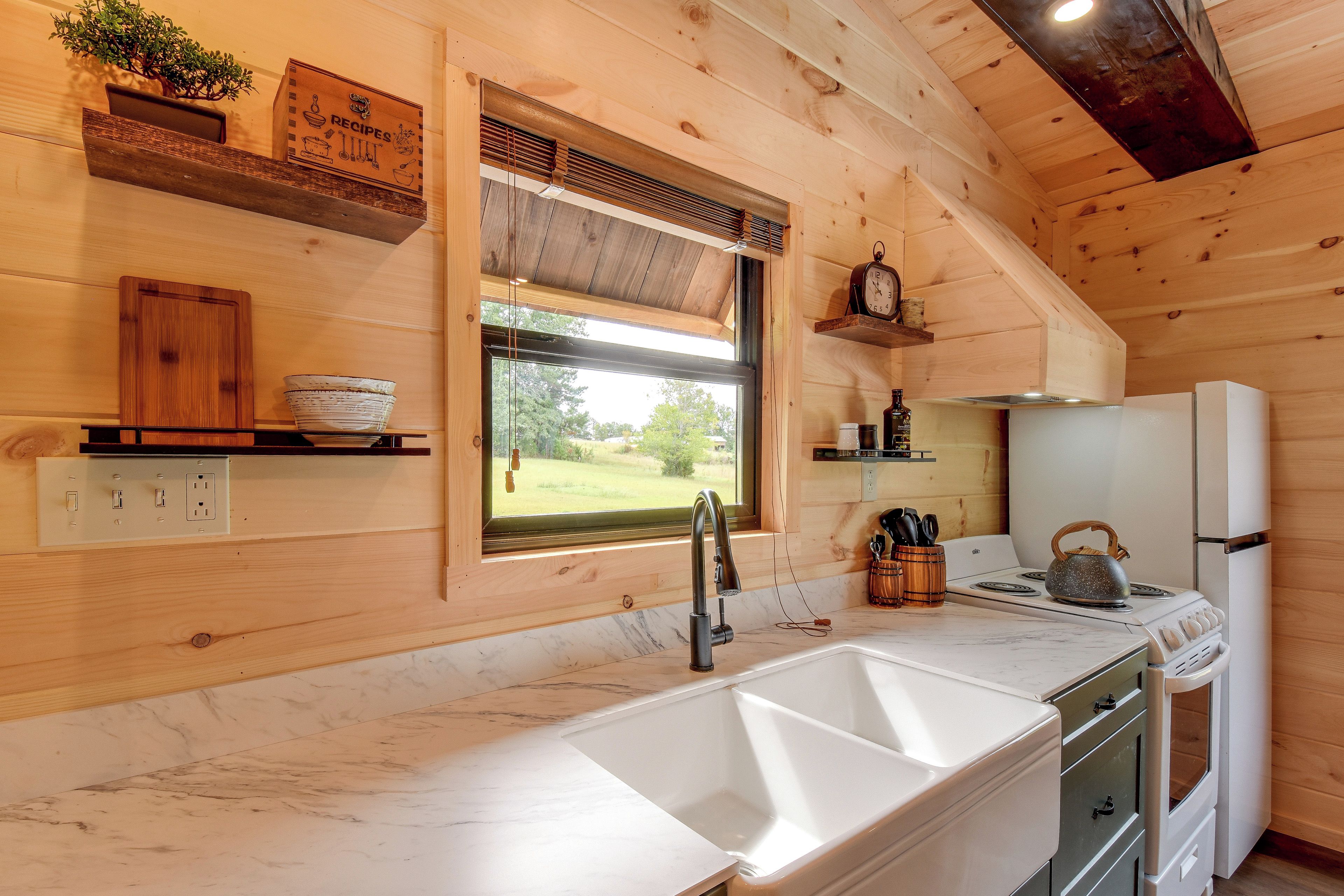 A kitchen with wooden walls, a marble countertop, a sink, a stove, and a window with a view of greenery.