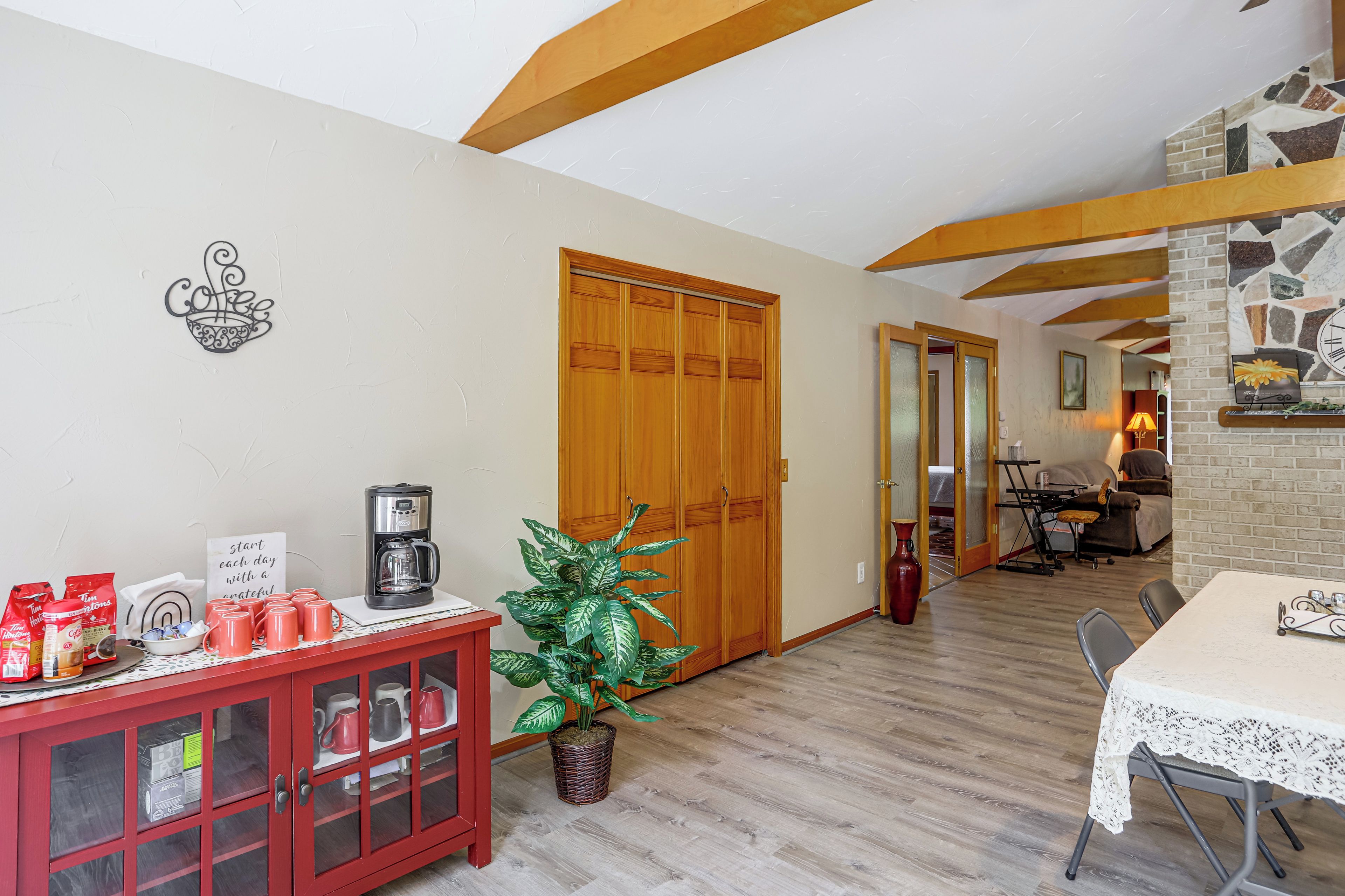 Una sala de estar acogedora con una estación de café, un mueble rojo con puertas de vidrio, una planta en maceta y una zona de comedor con una mesa y sillas.