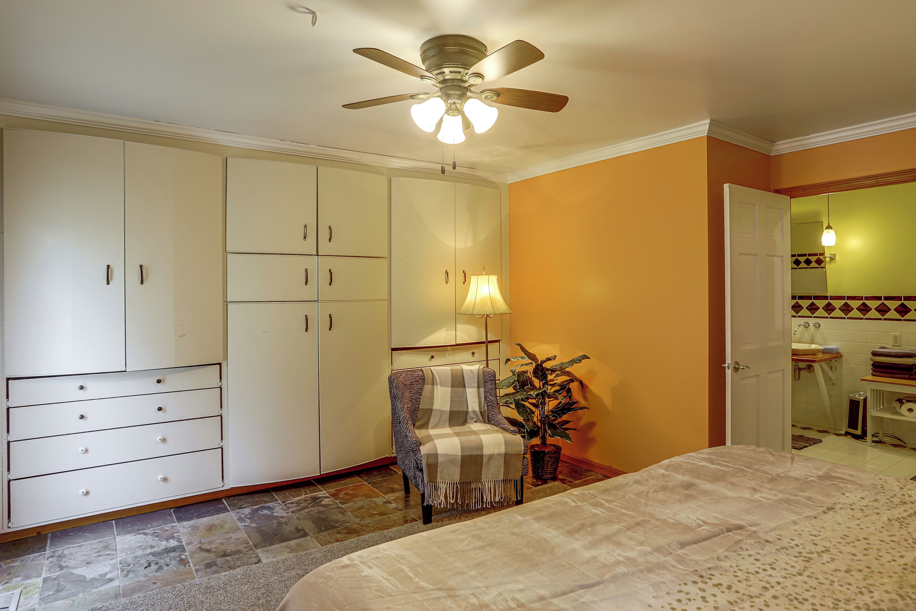 A bedroom with a bed, a chair, a ceiling fan, a lamp, and a bathroom visible through an open door.