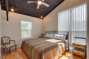 Un dormitorio con cama, una silla, una mesita de noche, un ventanal con persianas y un ventilador de techo.