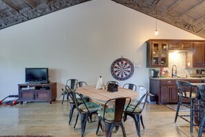 Un comedor con una mesa y sillas de madera, un dardo, un televisor en un mueble y una cocina con armarios de madera y una pared de piedra.