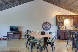Un comedor con una mesa y sillas de madera, un dardo, un televisor en un mueble y una cocina con armarios de madera y una pared de piedra.