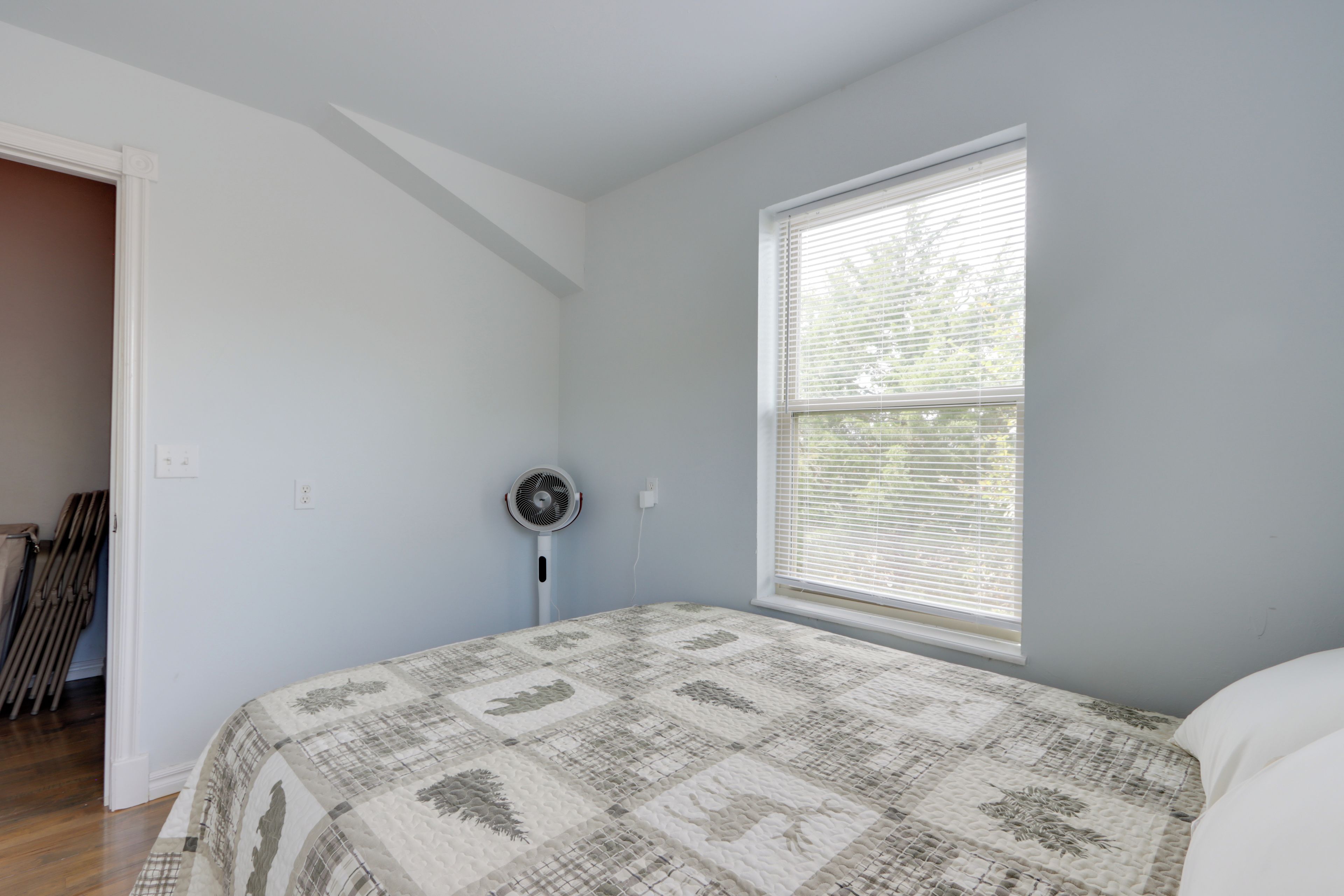 A bedroom with a bed, a standing fan, a window with blinds, and a door leading to another room.