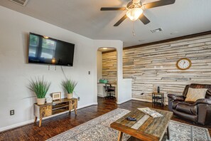 A living room with a flat-screen TV, a ceiling fan, a wooden wall, a clock, a leather sofa, and a coffee table. - Hot Tub & Patio! Family Escape, 7 Mi to Seaworld (San Antonio)