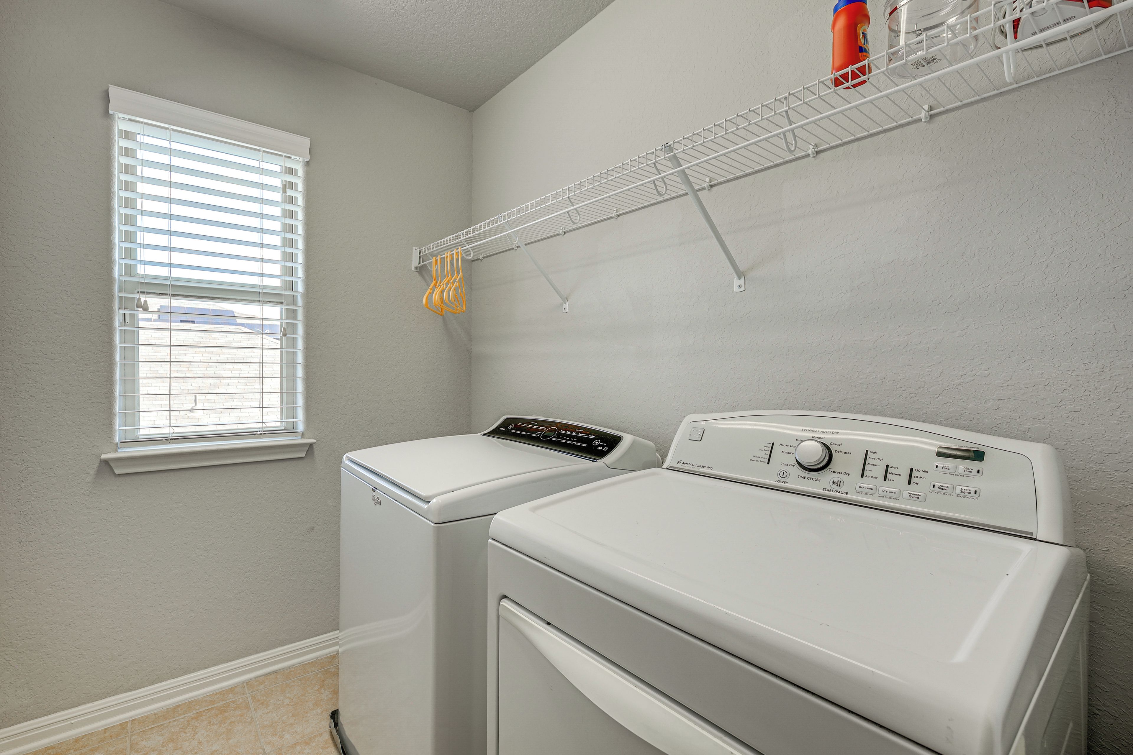 A laundry room with two white front-loading washing machines, a window with blinds, and a metal drying rack with hangers.