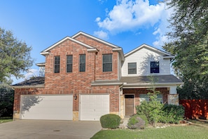 A two-story residential house with a brick facade, a white garage door, and a small front yard with landscaping. - Private Pool & Hot Tub: San Antonio Home (San Antonio)