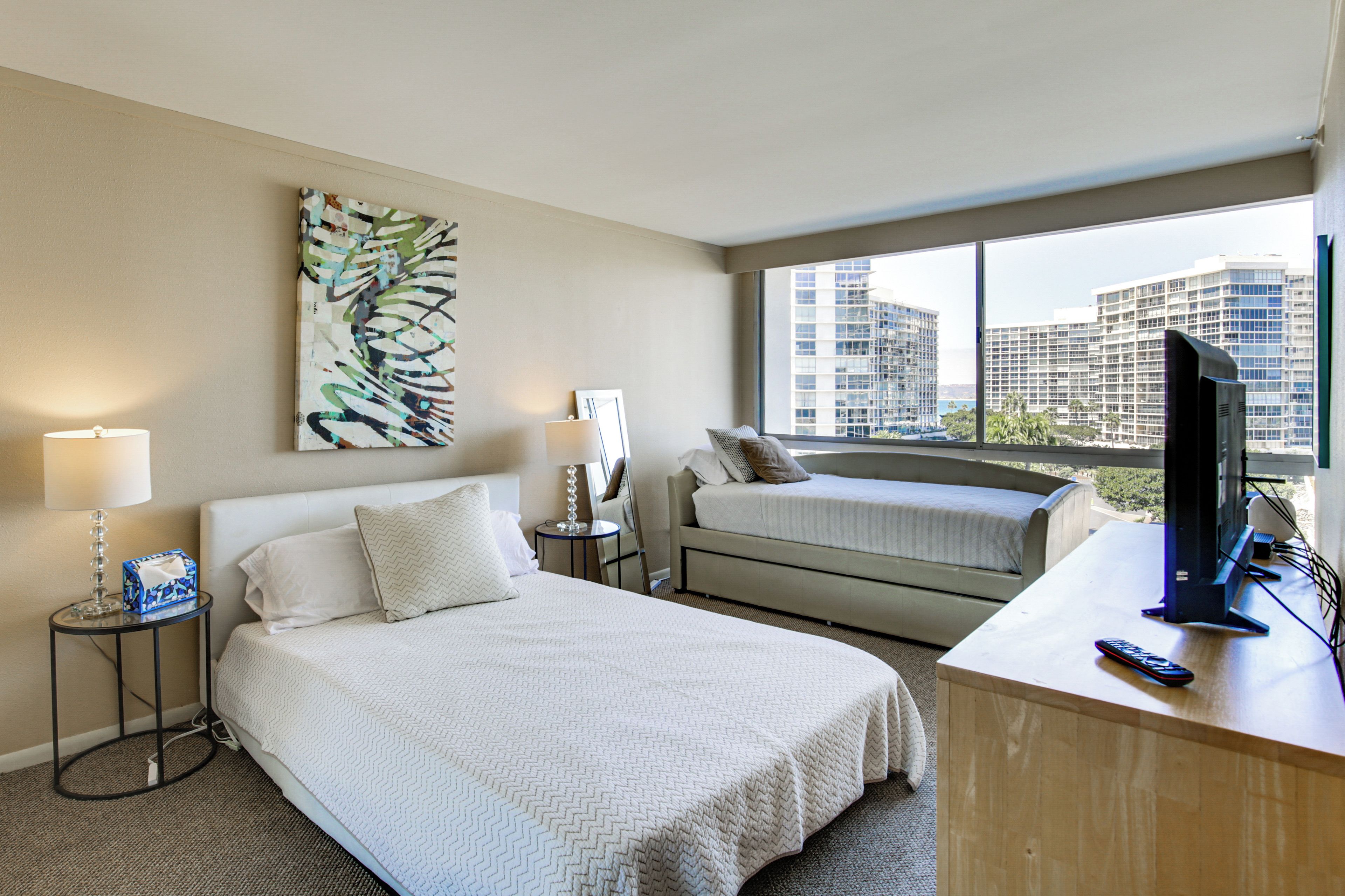 Quarto de hotel com cama, escrivaninha, computador, luminária e um quadro.
