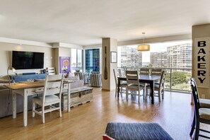 A modern living room with a flat-screen TV, a dining area with a table and chairs, and a large window with a view of high-rise buildings. - Beach + Poolside Days! Extended Coronado Escape (Coronado)