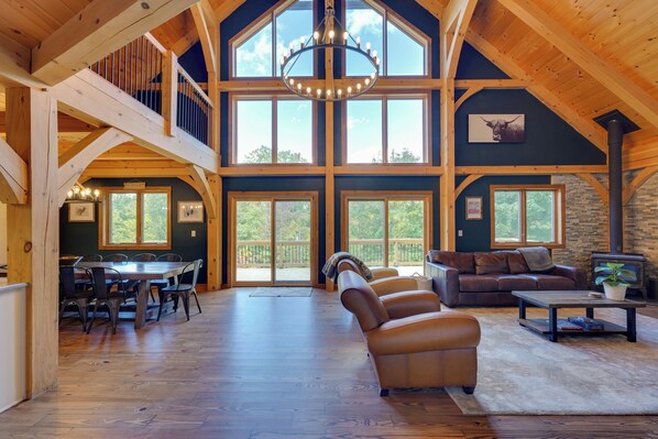 A spacious interior with a high ceiling, wooden beams, a fireplace, and a dining area with a view of the outdoors.