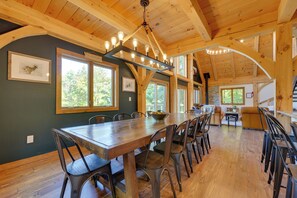 A spacious dining area with a long wooden table and chairs, large windows, and a high ceiling with exposed wooden beams.