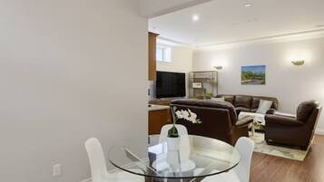 A modern living room with a glass dining table, white chairs, a brown sofa, and a flat-screen TV.