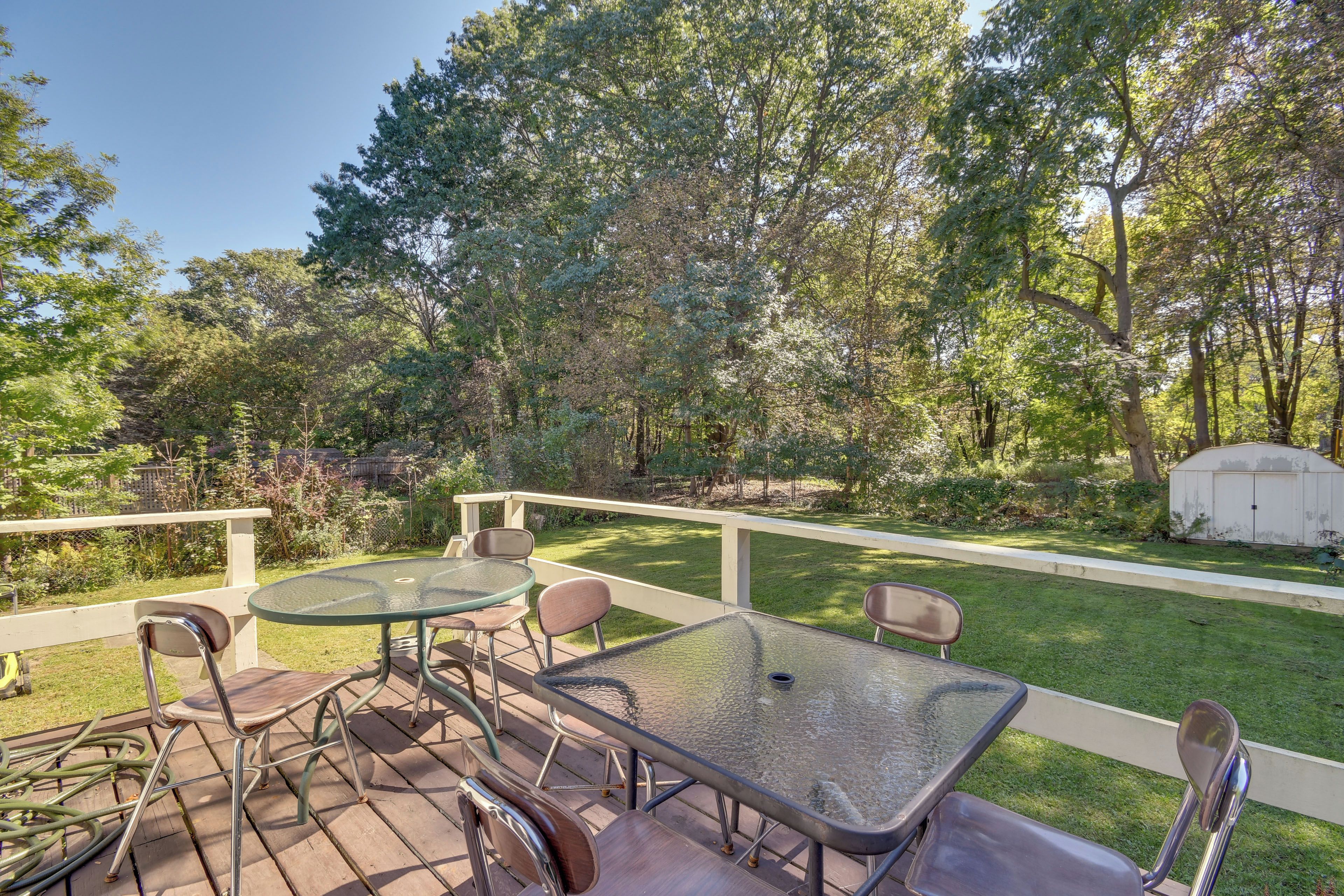 An outdoor patio with a glass table and chairs, surrounded by a white fence and lush greenery.