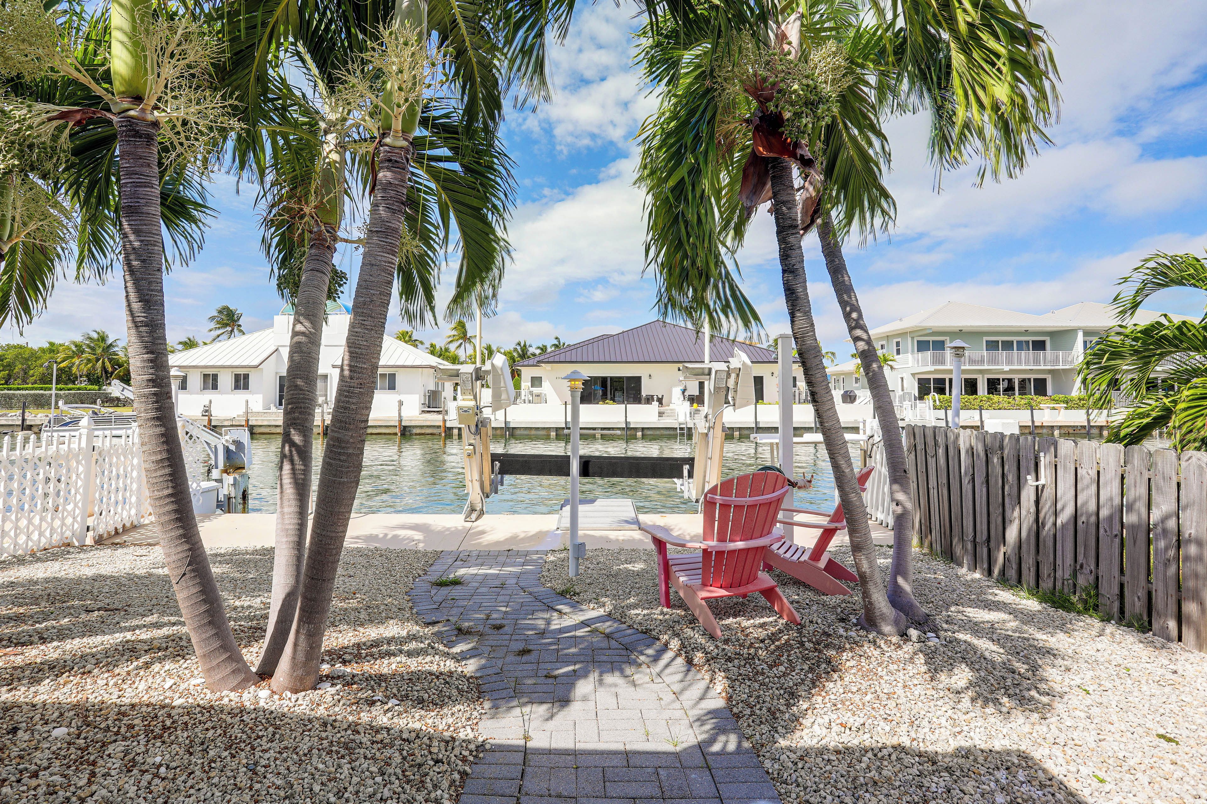 Propiedad frente al agua con muelle, palmeras, una silla Adirondack roja y un edificio con fachada blanca.