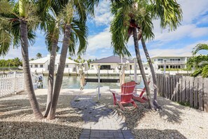 A waterfront property with a dock, palm trees, a red Adirondack chair, and a building with a white facade. - 1/2 Mi to Ocean! Canal-front Key Colony Beach Home (Key Colony Beach)
