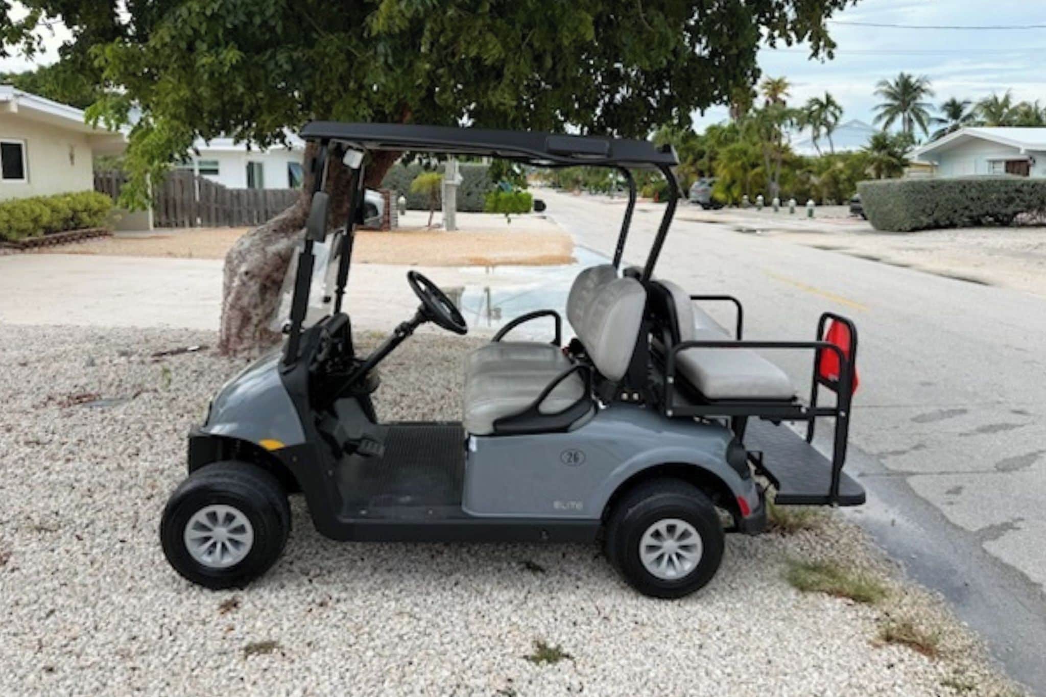 Un carrito de golf estacionado sobre grava, con un árbol y edificios residenciales al fondo.