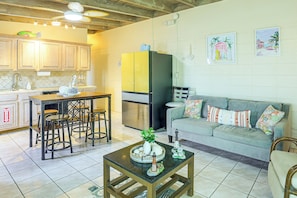 A compact living space with a kitchenette, a dining area with stools, a sofa with patterned cushions, and a small table with decorative items.