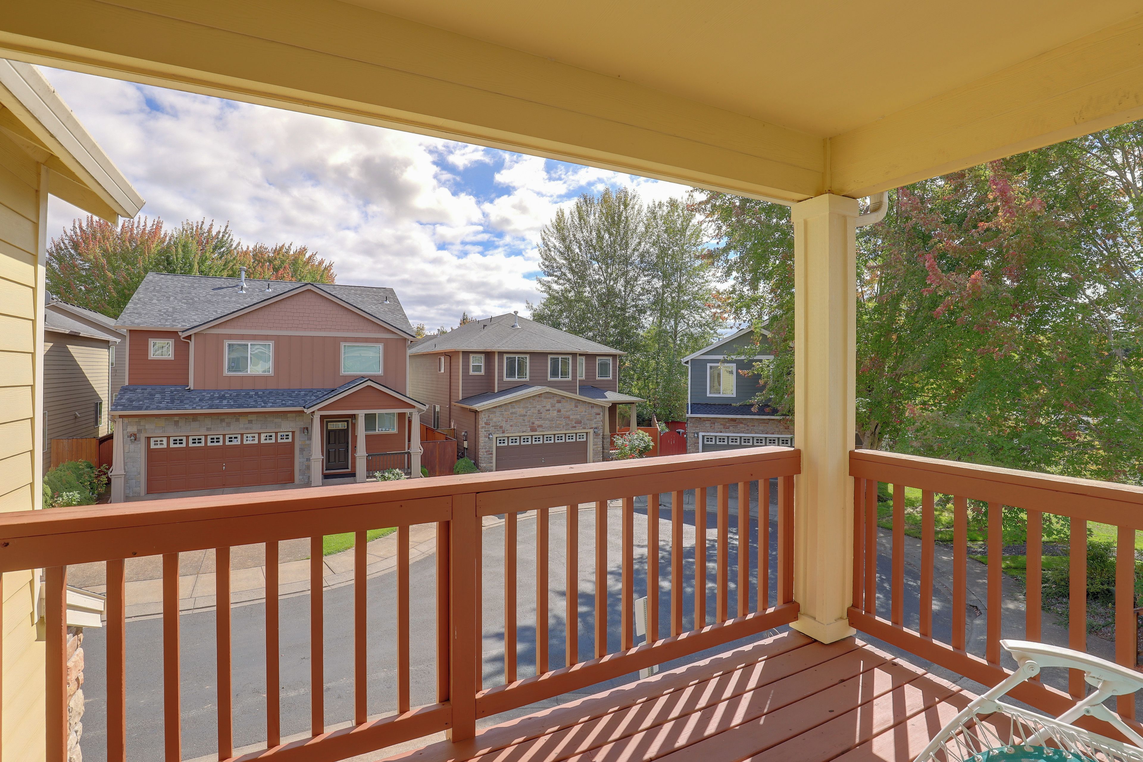 A covered porch with a wooden railing, overlooking a residential street with houses and a clear sky.
