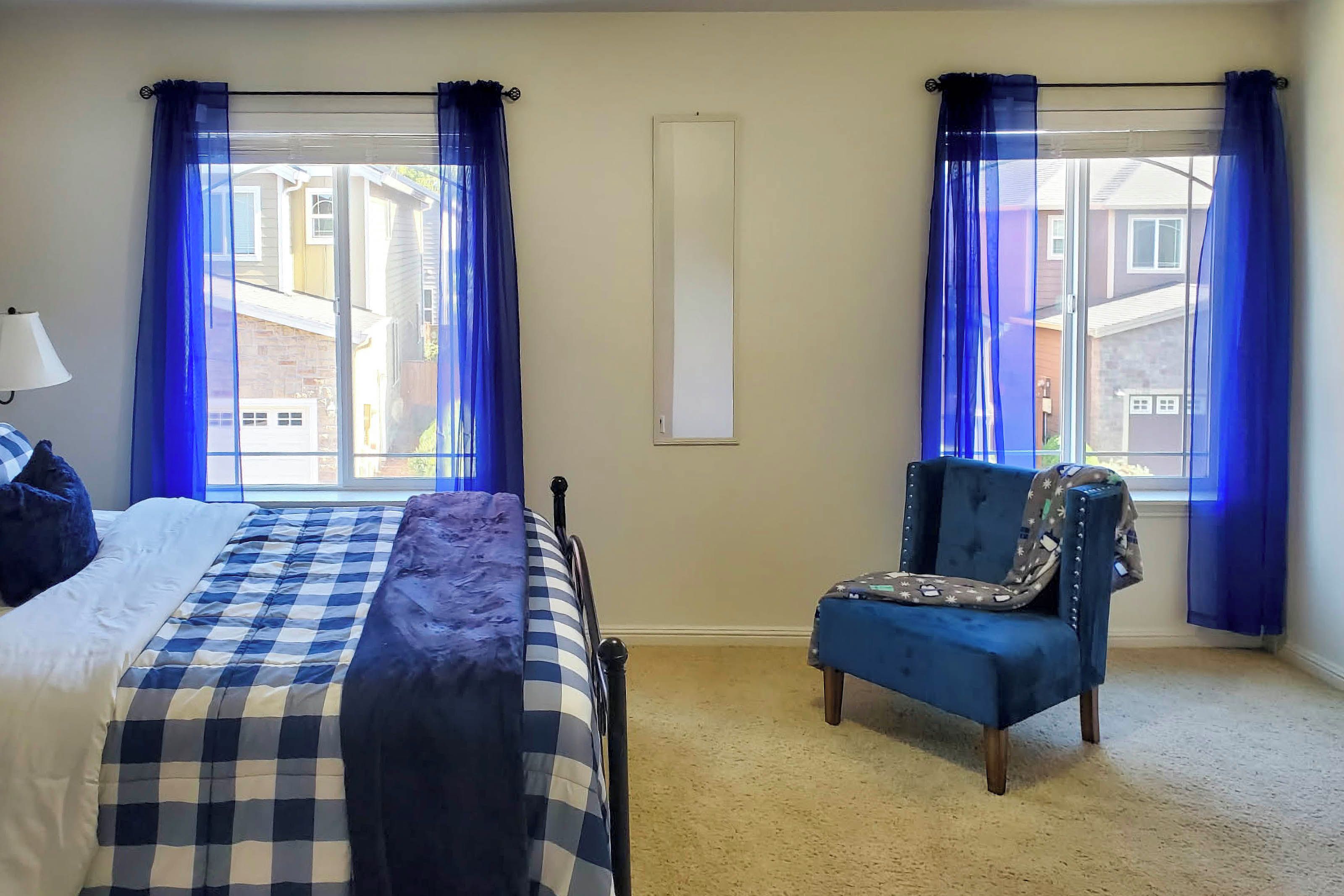 A bedroom with a bed, a chair, and two windows with curtains.