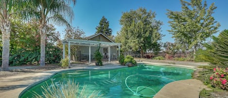 A backyard with a swimming pool, a covered patio, and various trees and plants.