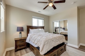 Quarto com cama, mesa de cabeceira, abajur, ventilador de teto e um espelho grande.
