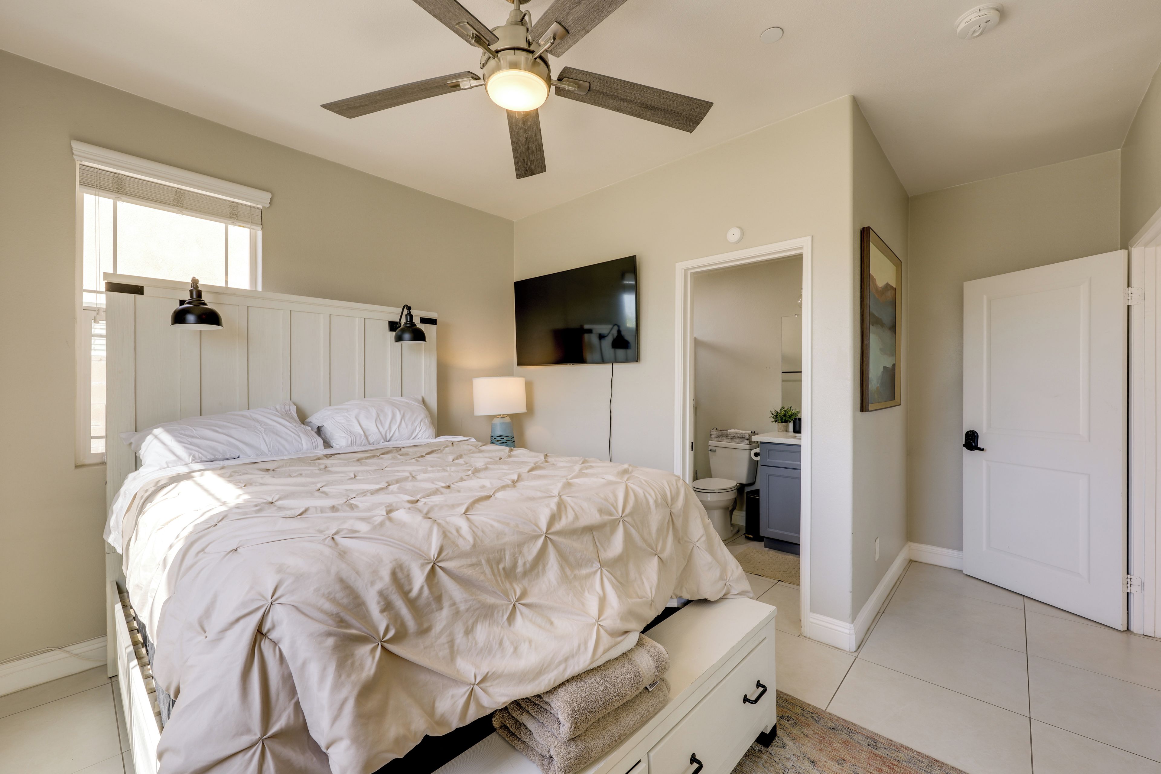 Une chambre à coucher comprenant un lit, un ventilateur de plafond, une télévision, une salle de bain avec des toilettes et un lavabo, ainsi qu’une porte.