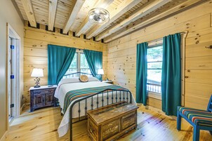 A wooden cabin bedroom with a bed, two bedside tables, a bench, a chair, and a window with curtains.
