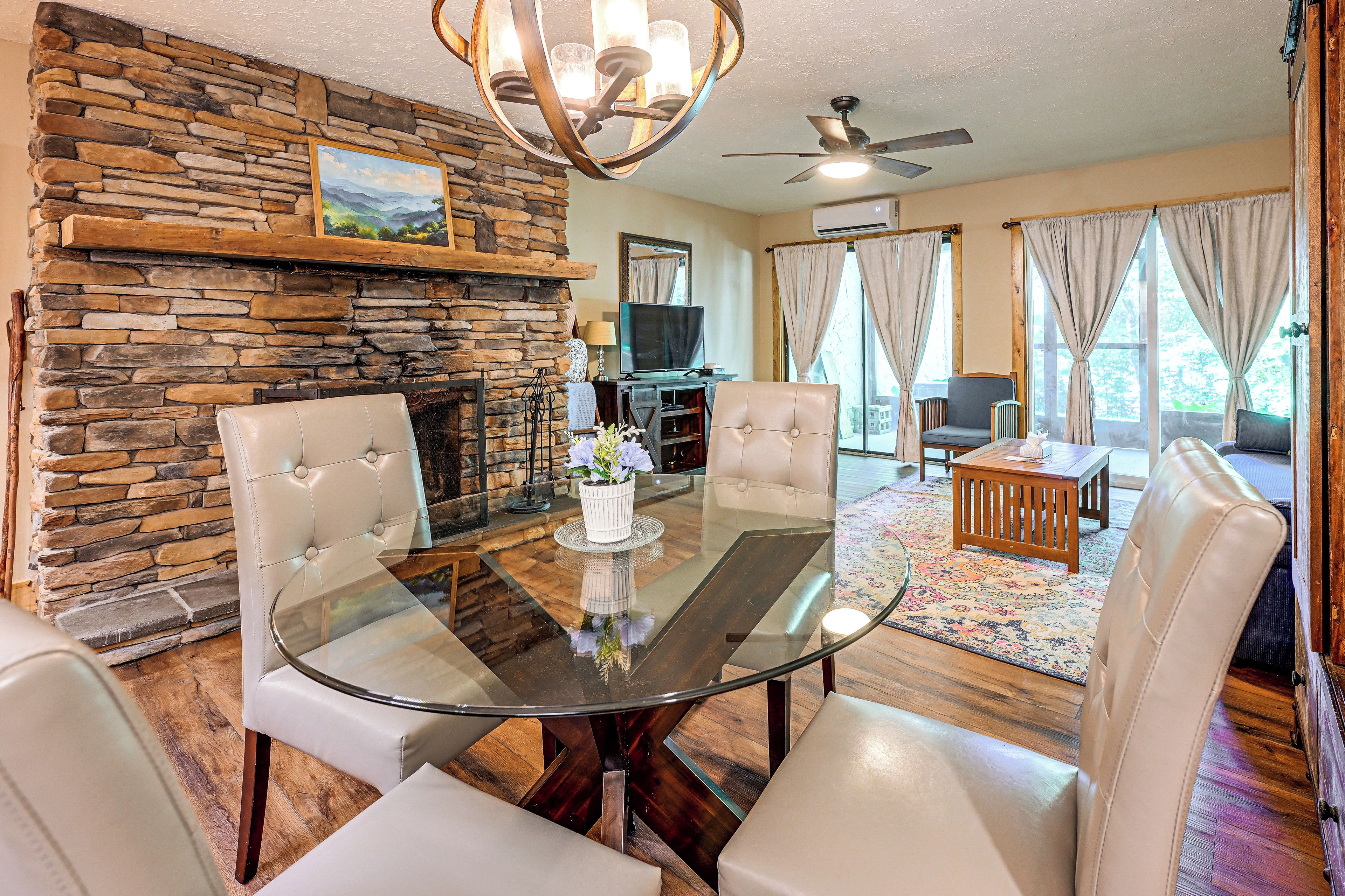 A living room with a stone fireplace, a glass dining table, leather chairs, a ceiling fan, and a television.
