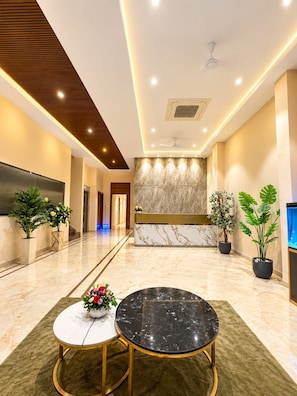 Um saguão de hotel com balcão de recepção, piso de mármore, plantas em vasos e uma tela grande.