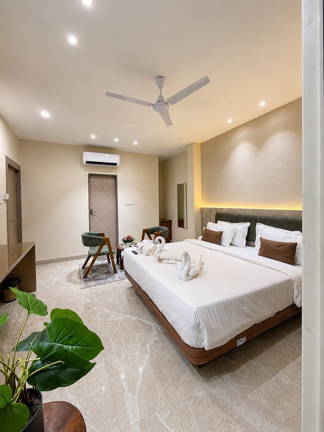 A hotel room with a large bed, a ceiling fan, an air conditioning unit, a desk, a chair, and a potted plant.