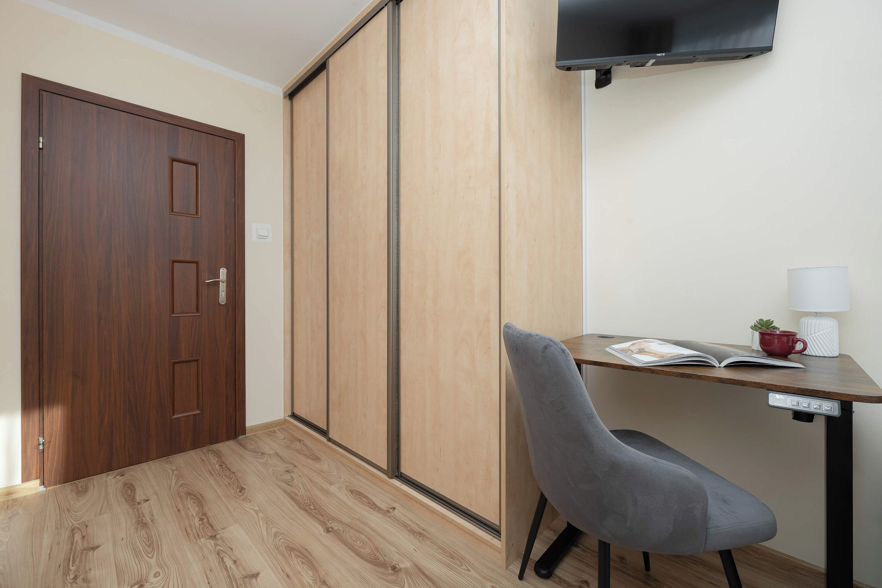 This compact room offers a relaxing atmosphere with its neatly made bed, a wall-mounted TV, and a dedicated workspace.